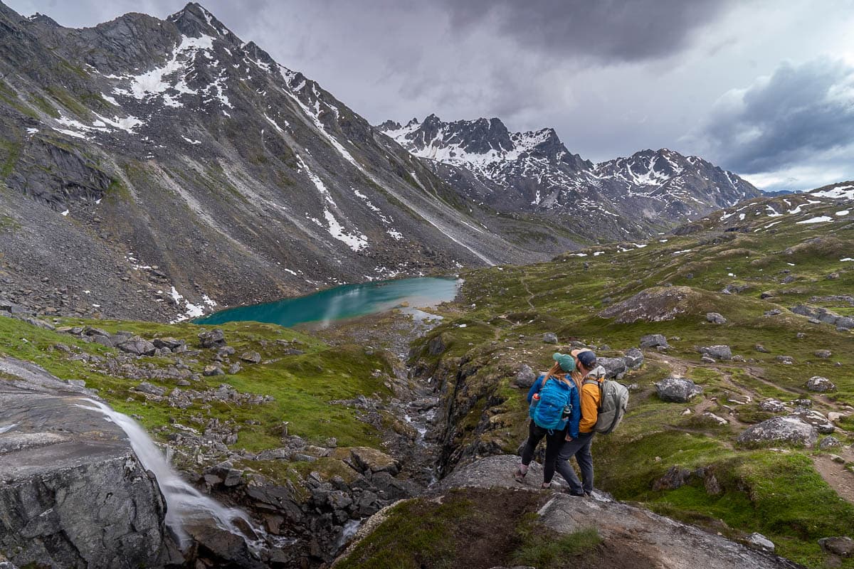 Couple standing on a rocky outcropping overlooking Reed Lake with the Talkeetna Mountains in the background along the Reed Lakes Trail in Hatcher Pass, Alaska