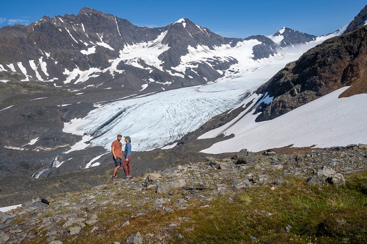 Couple standing in front of the Raven Glacier surrounded by Chugach Mountains along the Crow Pass Trail in Chugach Mountain State Park in Anchorage, Alaska