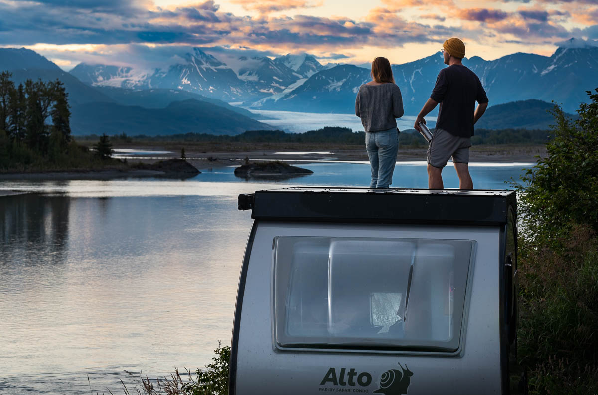 Couple standing on top of a Safari Condo Alto F1743 Expedition trailer with the Knik Glacier surrounded by the Chugach Mountains in Palmer, Alaska
