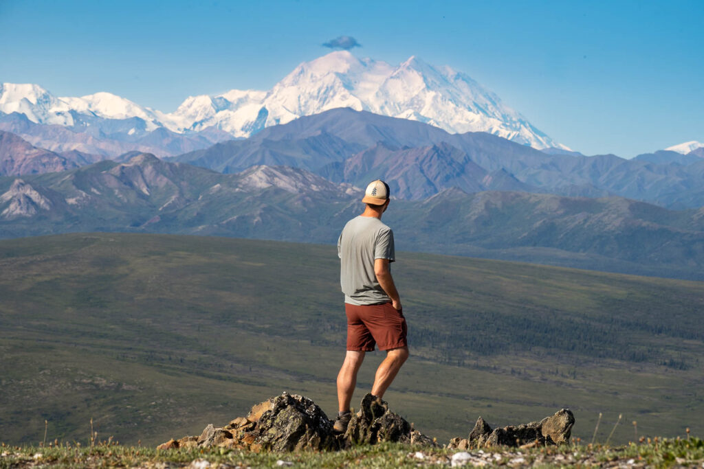 Man standing on a rocky outcropping with Mount Denali in the background along the Savage Alpine Trail in Denali National Park, Alaska