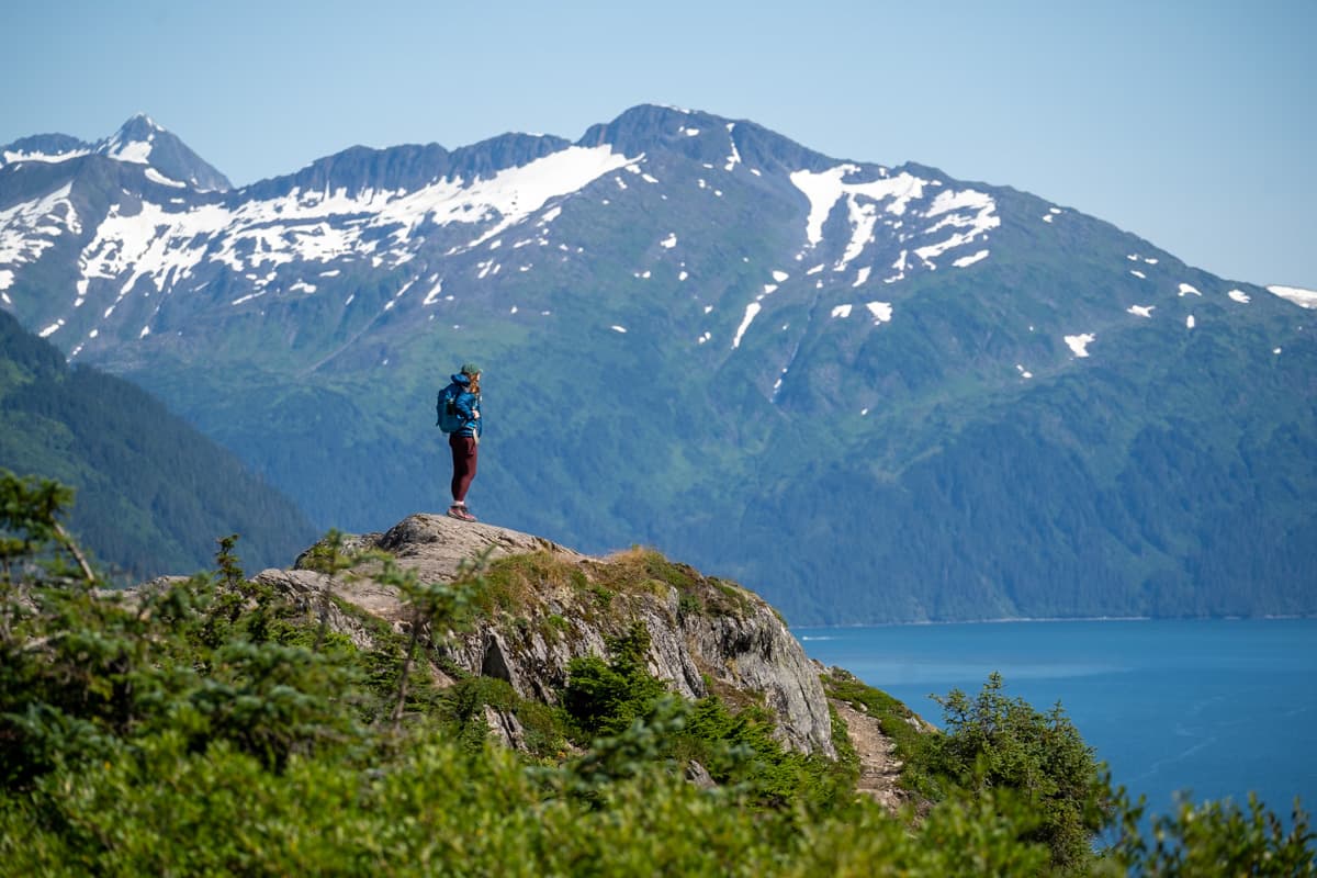 Woman standing on a rocky outcropping with the Chugach Mountains and Prince William Sound in the background along the Portage Pass Trail in Whittier, Alaska