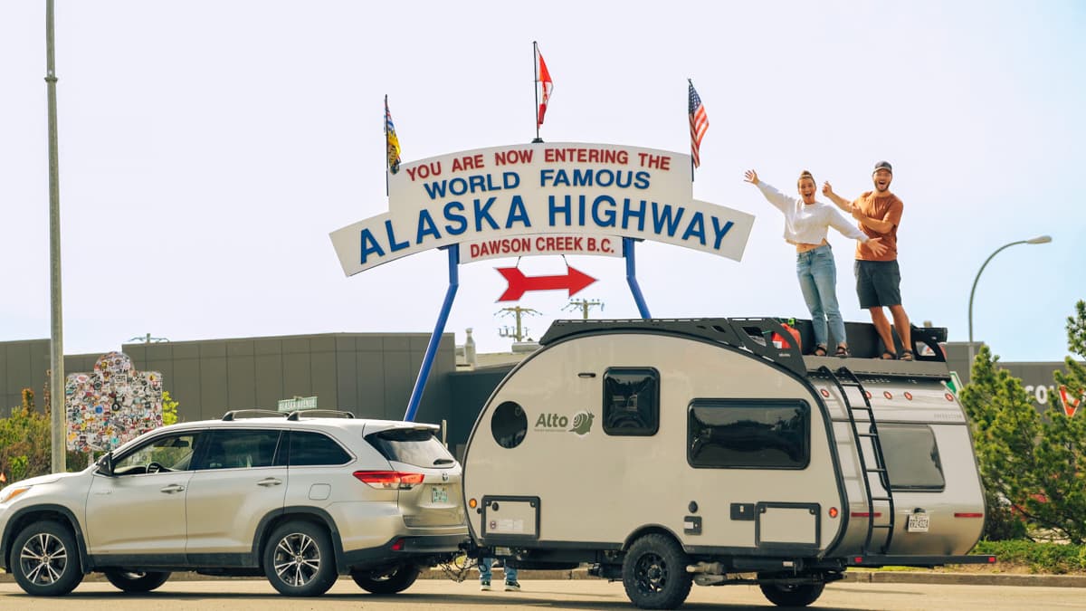 Couple standing on top of the Safari Condo Alto F1743 Expedition with the Alaska Highway sign in the background in Dawson Creek, British Columbia, Canada