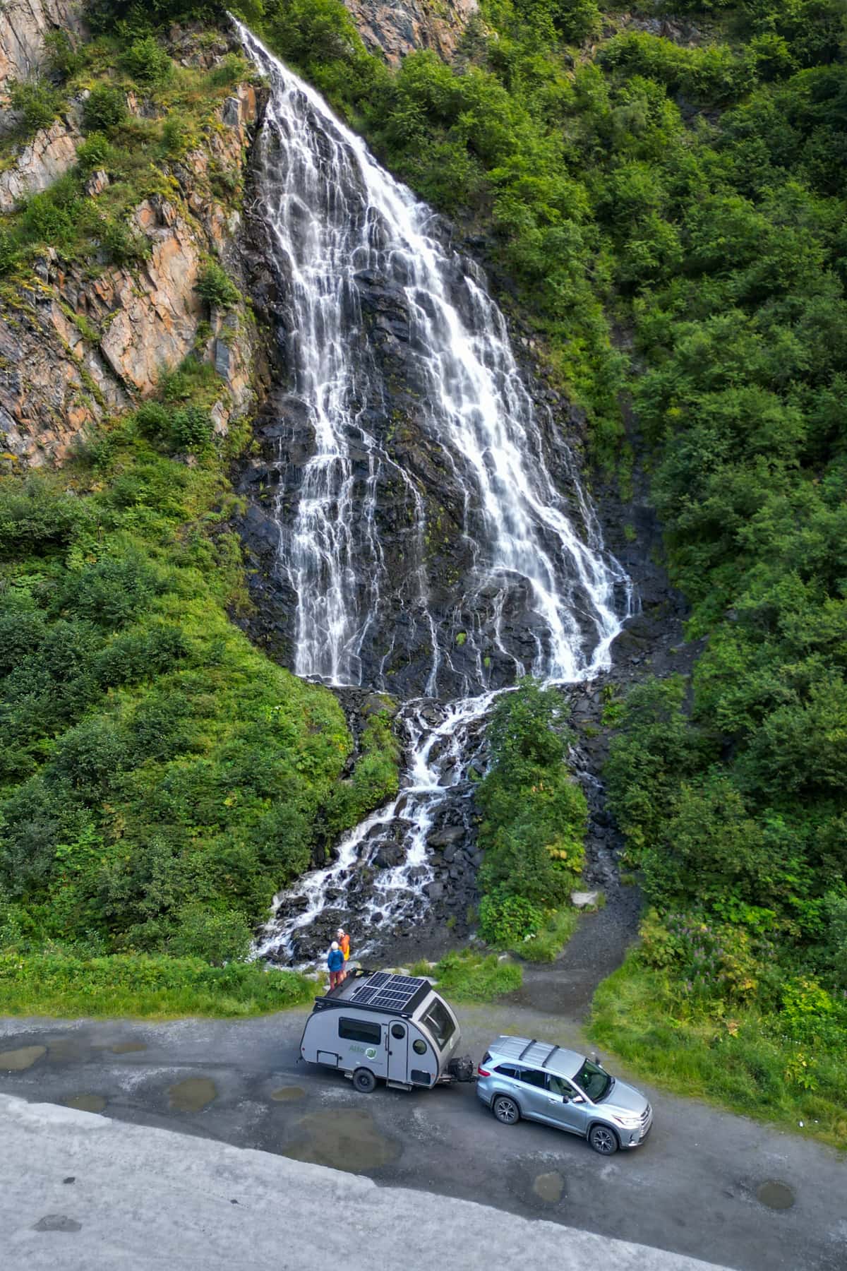 Couple standing on top of a Safari Condo Alto F1743 Expedition trailer with Horsetail Falls in the background in Keystone Canyon of Valdez, Alaska