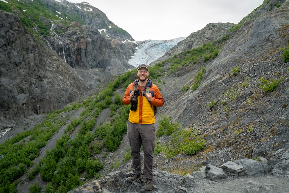Man standing on a rocky outcropping with the Exit Glacier and the Kenai Mountains in the background along the Exit Glacier Trail in Kenai Fjords National Park, Alaska