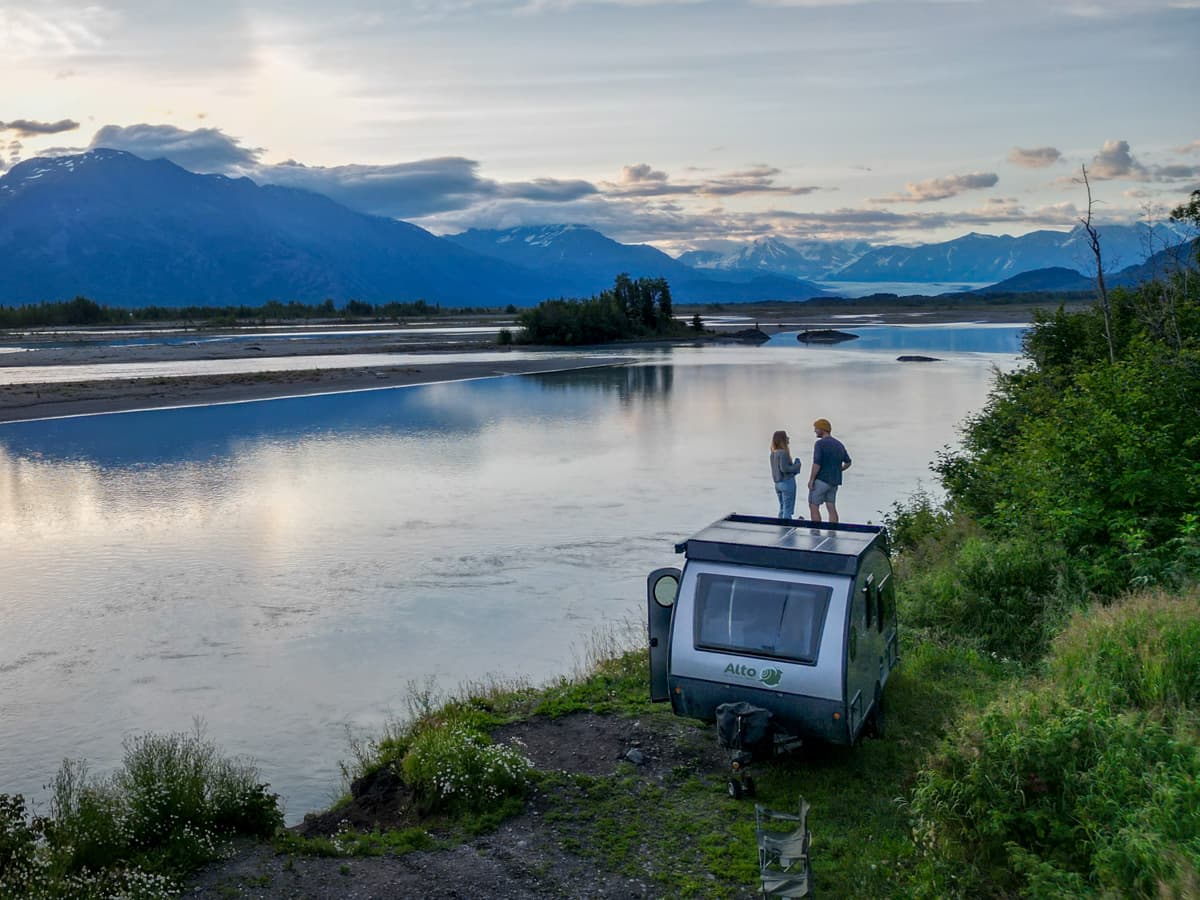 Couple standing on top of a Safari Condo Alto F1743 Expedition trailer with mountains and the Knik Glacier in the background in Palmer, Alaskae