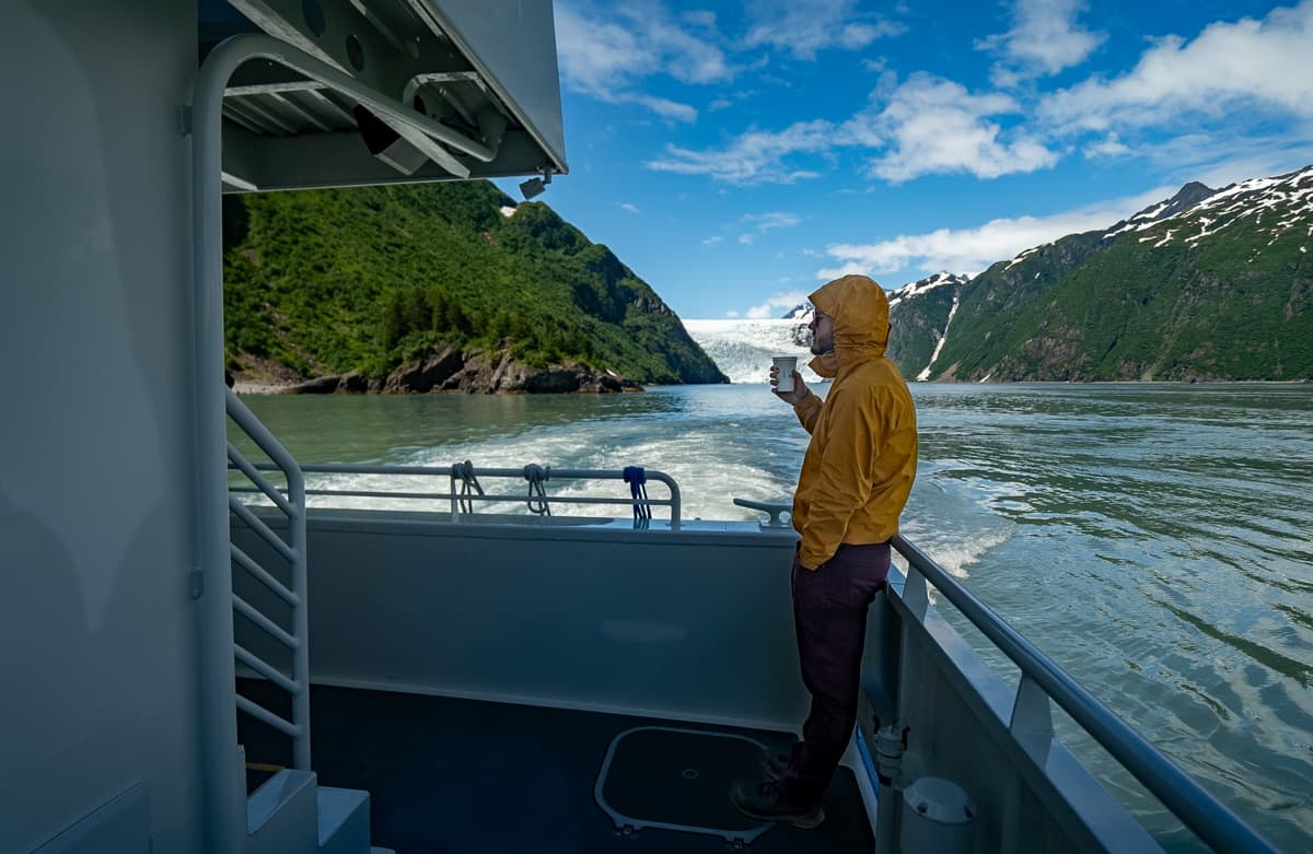 Man drinking out of a cup on a Major Marine cruise ship with a glacier and mountains in the background in Kenai Fjords National Park, Alaska