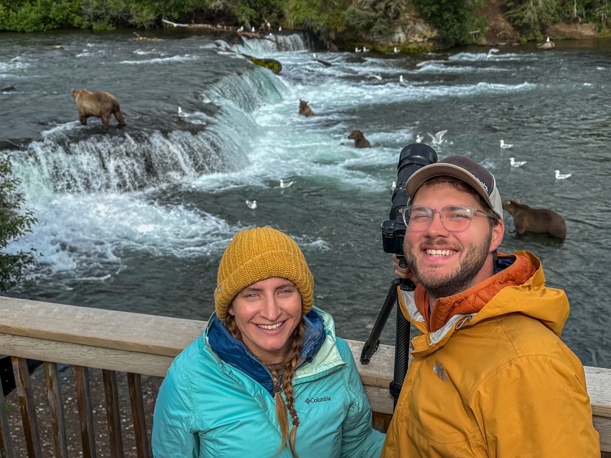 Smiling couple standing on a wooden platform with bears fishing on Brooks Falls in the background in Katmai National Park, Alaska