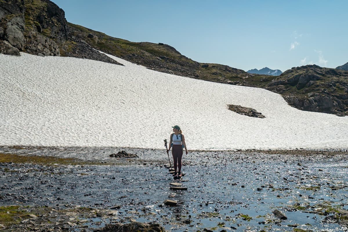 Woman standing on a rock along a flooded trail with a snowy mountain in the background along the Crow Pass Trail in Chugach State Park, Alaska