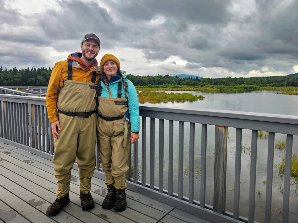 Smiling couple wearing waders while standing on a wooden boardwalk at Brooks Camp in Katmai National Park, Alaska