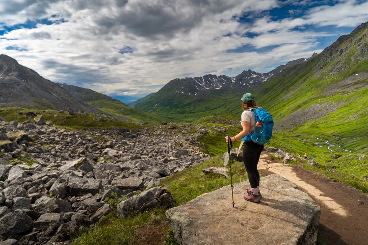 Woman with trekking poles standing on a rocky outcropping with the Talkeetna Mountains in the background along the Reed Lakes Trail in Hatcher Pass, Alaska