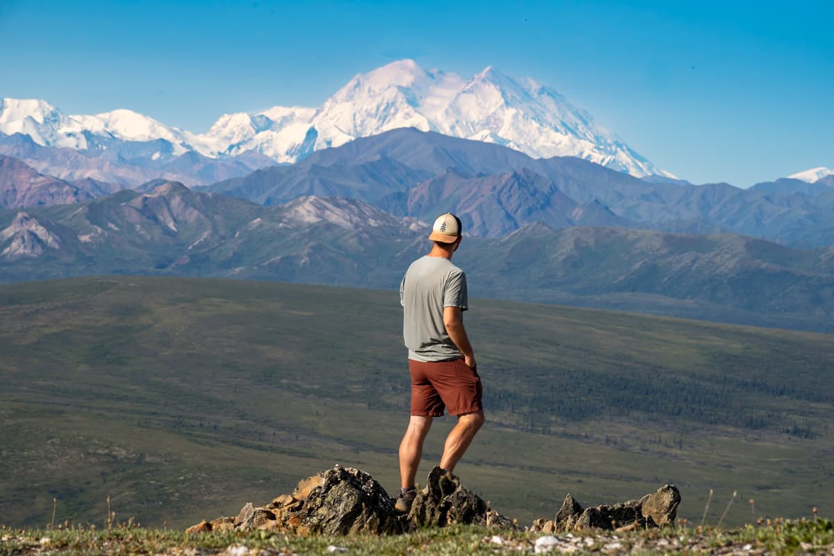 Man standing on a rocky outcropping with Mount Denali and the Alaska Mountain range in the background along the Savage Alpine Trail in Denali National Park, Alaska