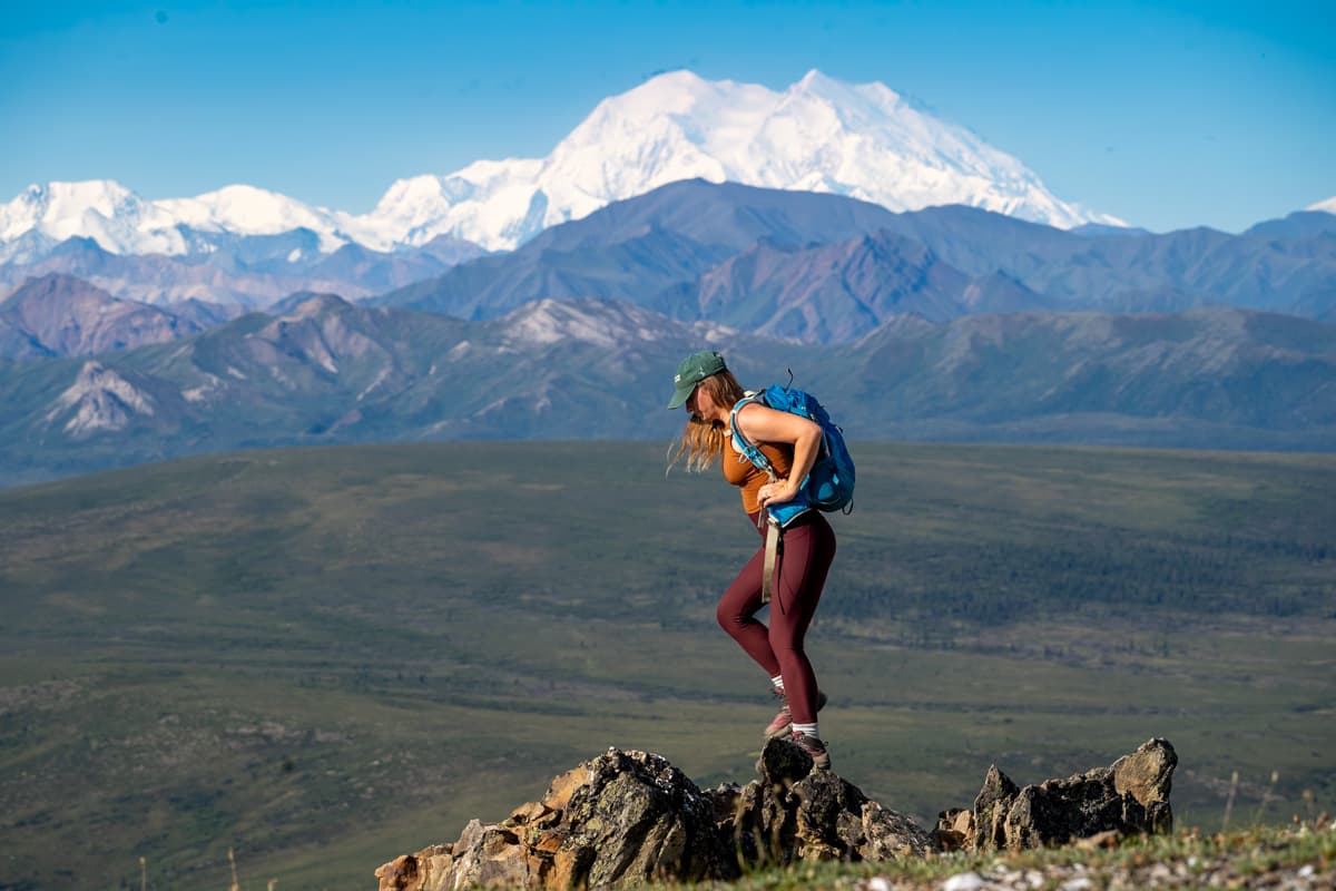 Woman standing on a rocky outcropping with Mount Denali and the Alaska Mountain range in the background along the Savage Alpine Trail in Denali National Park in Alaska
