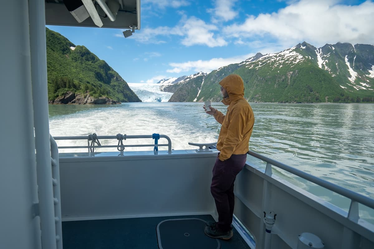 Man standing at the railing of a boat with mountains and glaciers in the background on a Major Marine tour in Kenai Fjords National Park in Alaska