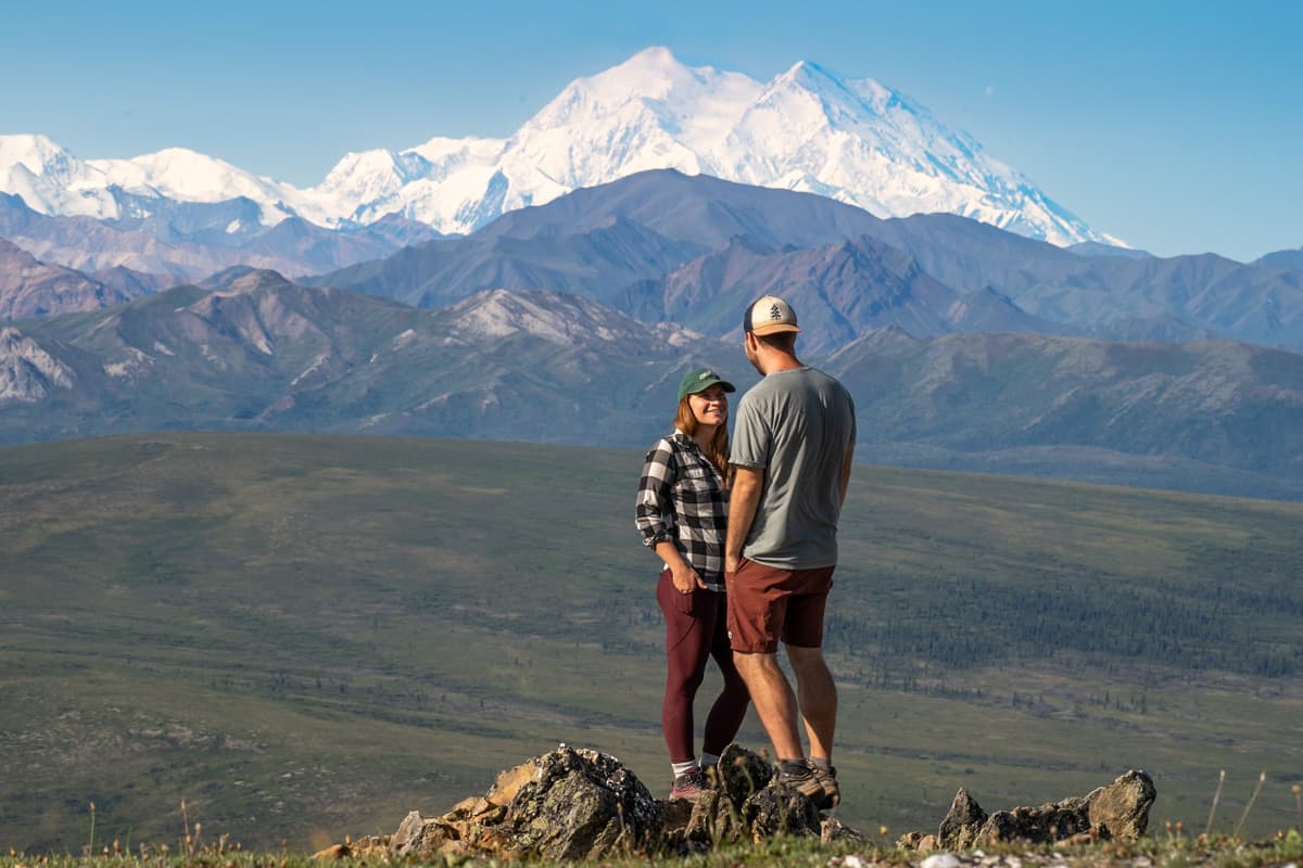 couple standing on a rocky outcropping with Mount Denali and the Alaska Mountain range in the background along the Savage Alpine Trail in Denali National Park in Alaska