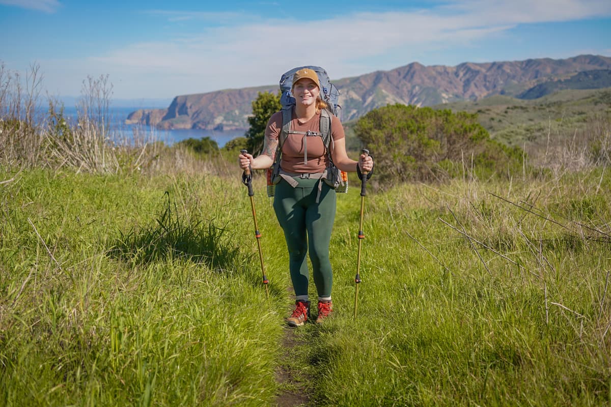 Woman wearing a backpack and trekking poles along the Del Norte Trail with cliffsides in the background on Santa Cruz Island in Channel Islands National Park, California