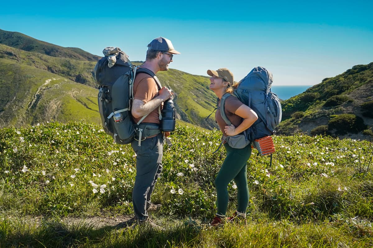 Smiling couple wearing backpacks standing next to a field of wildflowers with rolling green hills in the background on Santa Cruz Island in the Channel Islands National Park, California