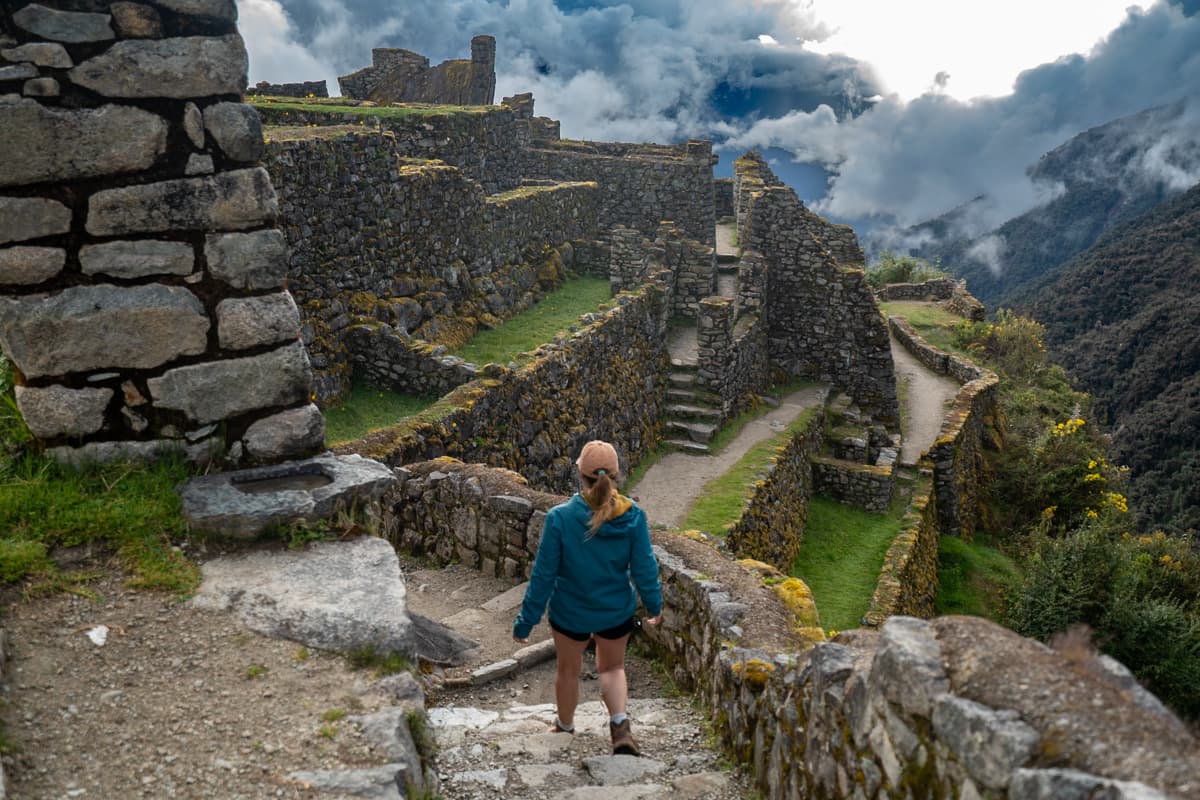 Woman walking down the stairs at Sayacmarca Inca ruins with mountains and low-hanging clouds in the background along the Inca Trail in Peru