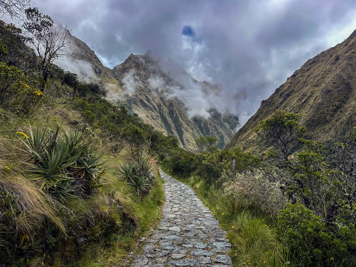 Stone pathway of the Inca Trail leading through green mountains with low hanging clouds in the Andes Mountains of Peru