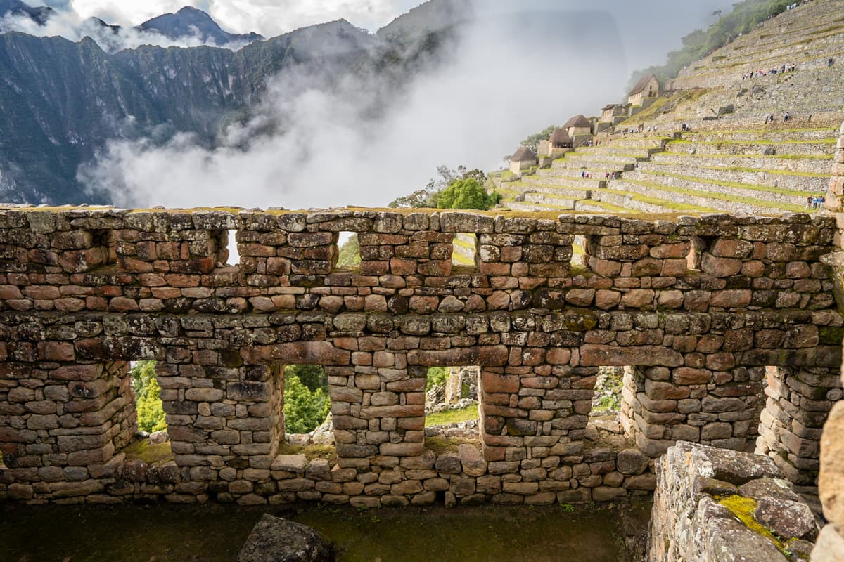 Stone wall with windows with low-hanging clouds and mountains in the background in Machu Picchu, Peru