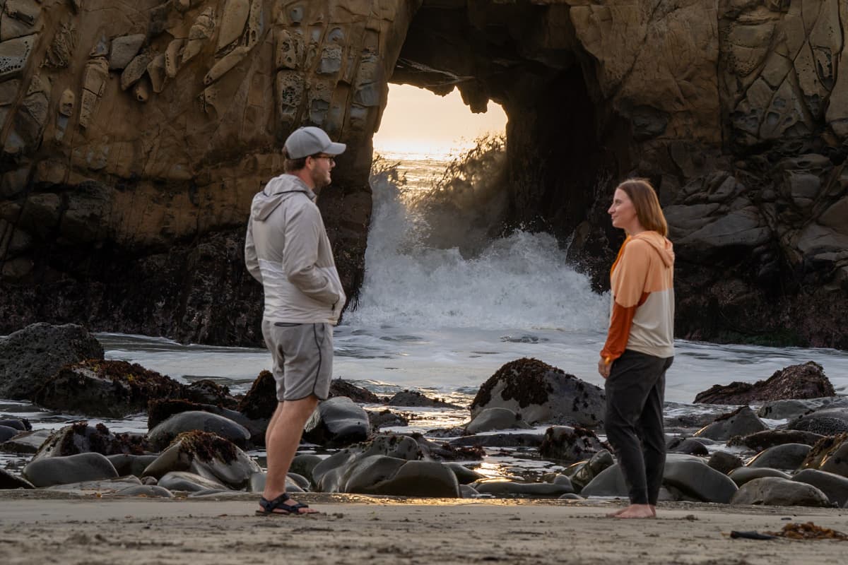 Couple standing in front of Keyhole Arch with waves crashing through it at Pfeiffer Beach in Big Sur, California