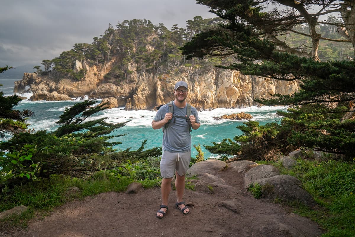 Smiling man standing in front of a cliffside with turquoise water and rock formations in the background along the Cypress Grove Trail in Point Lobos State Natural Reserve in Big Sur, California