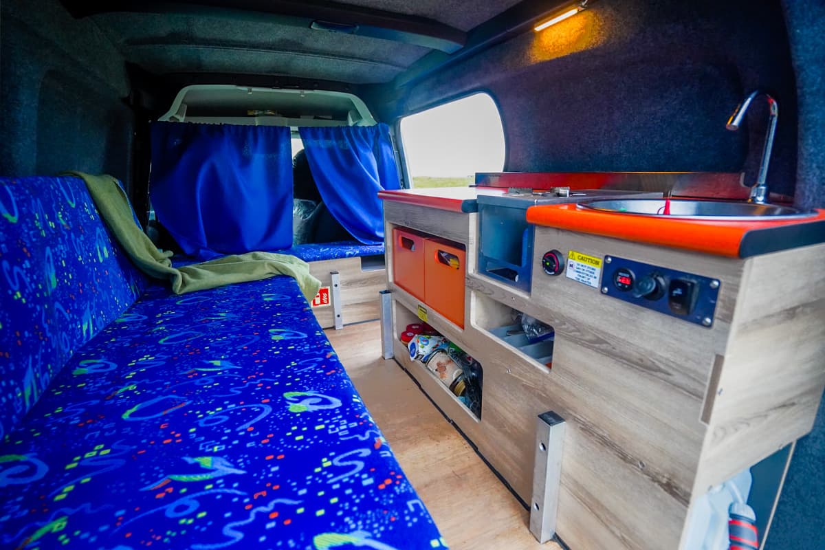Interior shot of a couch and kitchen setup in the Happy 1 model of Happy Campers campervan in a grassy field in Iceland