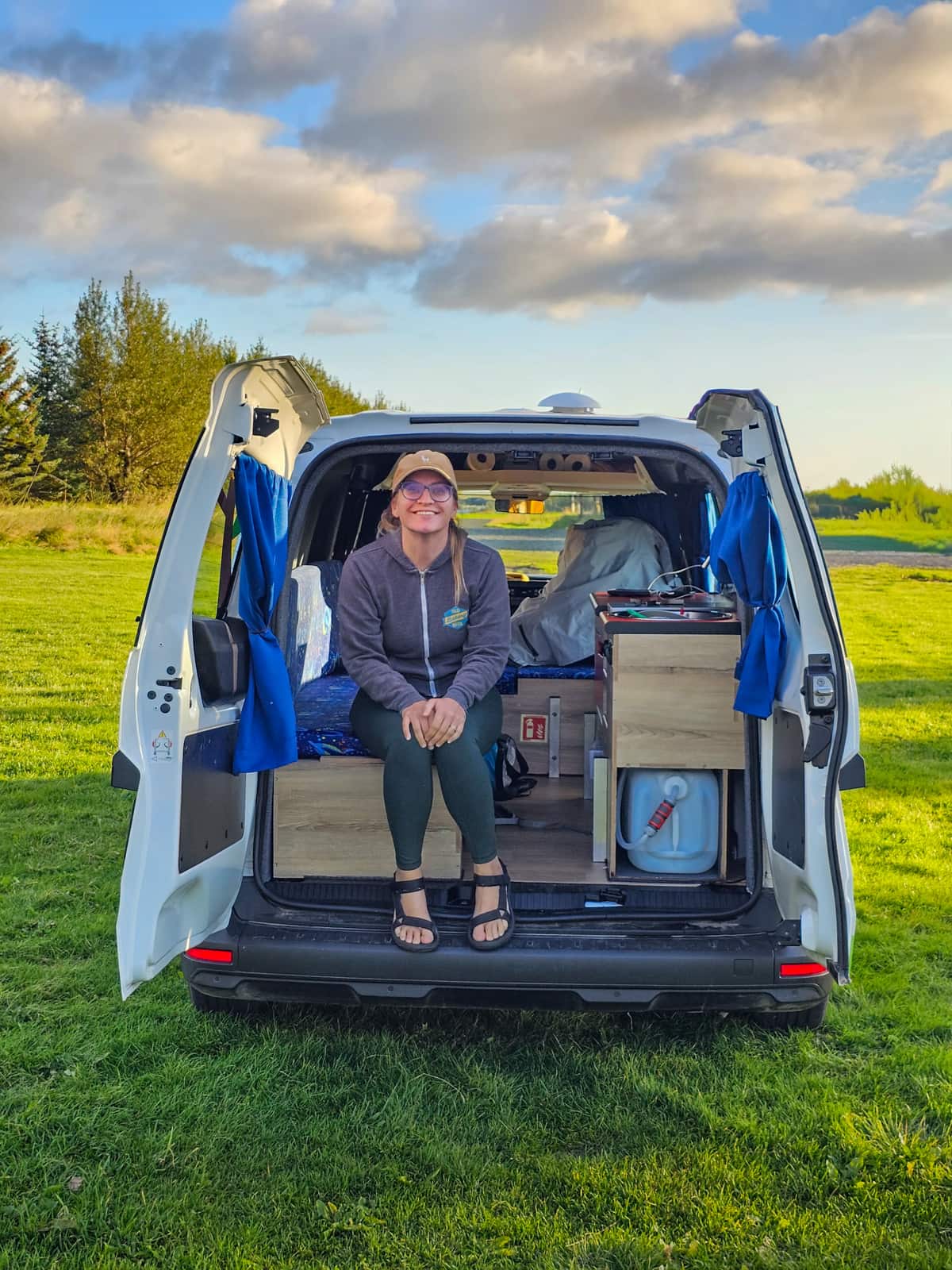 Smiling woman sitting in the back of a campervan with open doors while sitting in a grassy field in Iceland