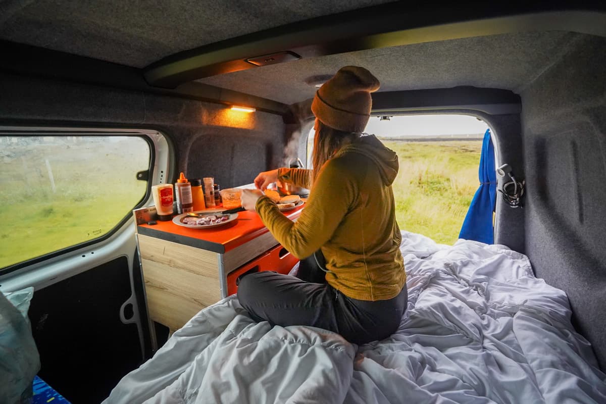 Woman sitting on her bed cooking in a campervan with a grassy field in the background in Iceland