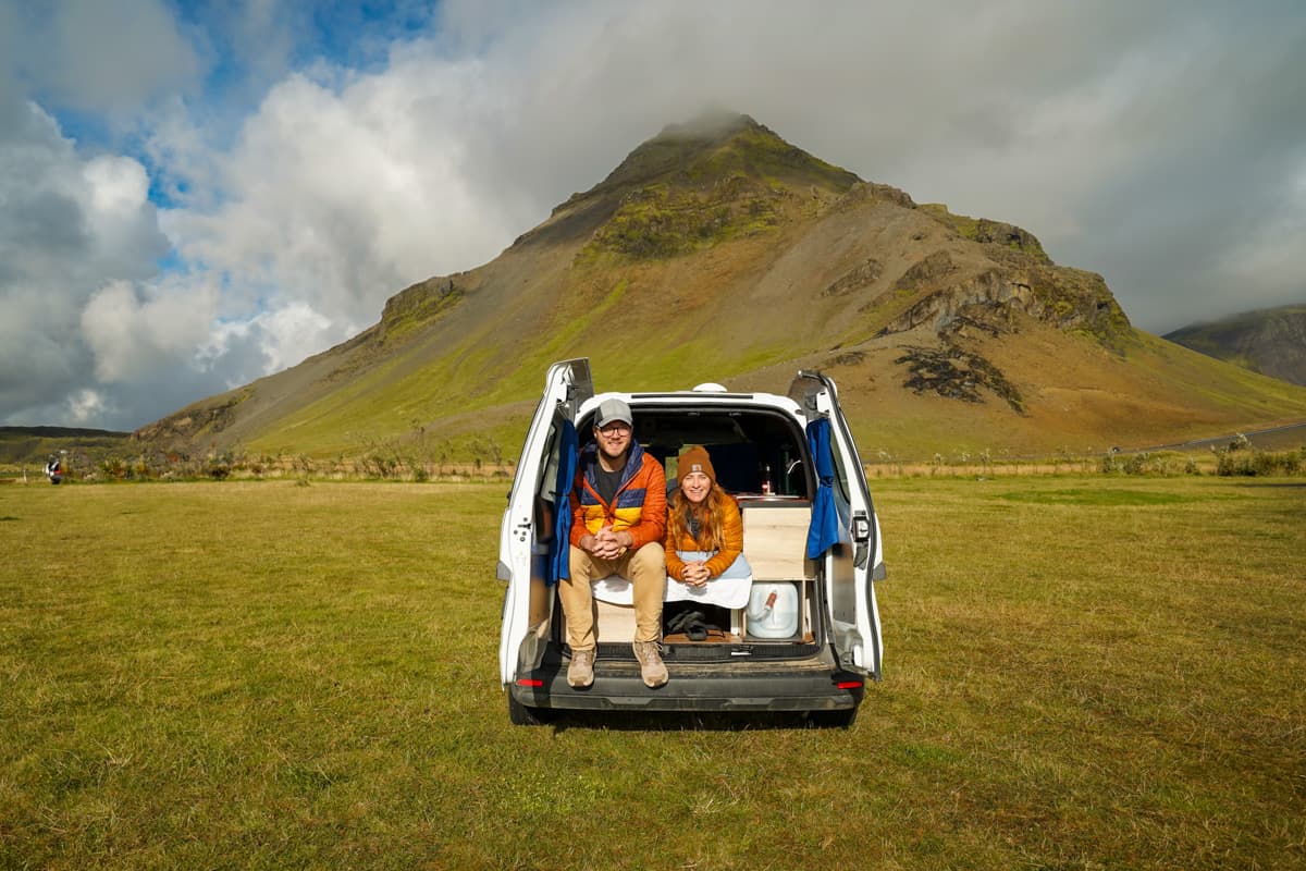 Couple sitting on a bed with the doors open on their Happy Campers campervan with Stapafell mountain in the background in Iceland