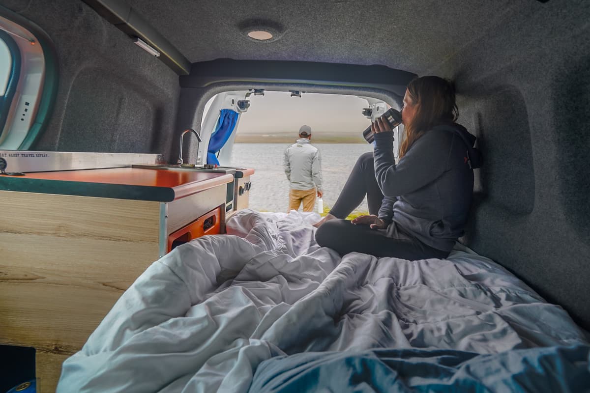 Woman sitting on a bed in a campervan drinking coffee while a man stands in the background of open doors, overlooking a fjord, in Iceland