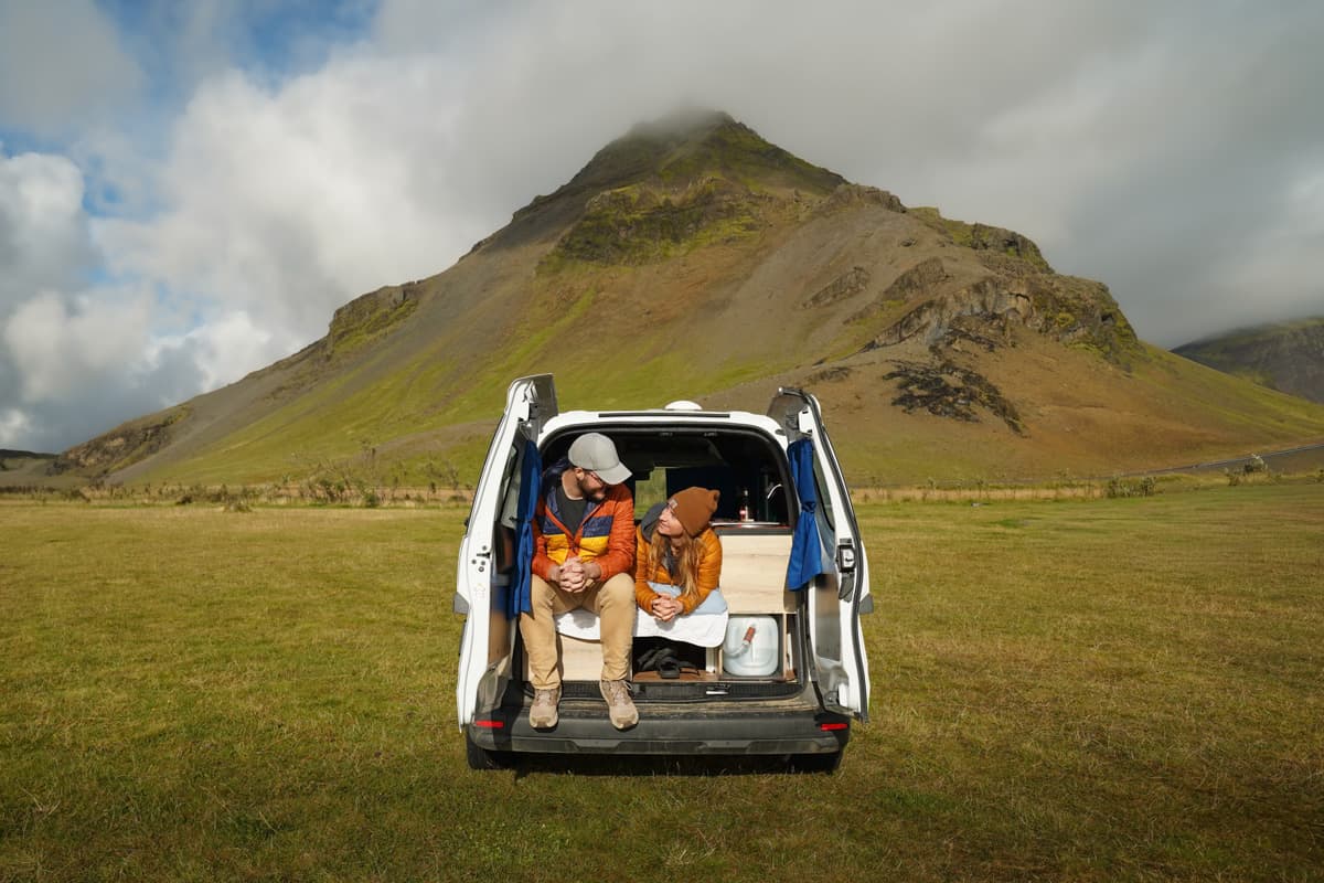 Couple laying on the bed in the back of a campervan with open doors with Stapafell mountain in the background in Arnastapi, Iceland
