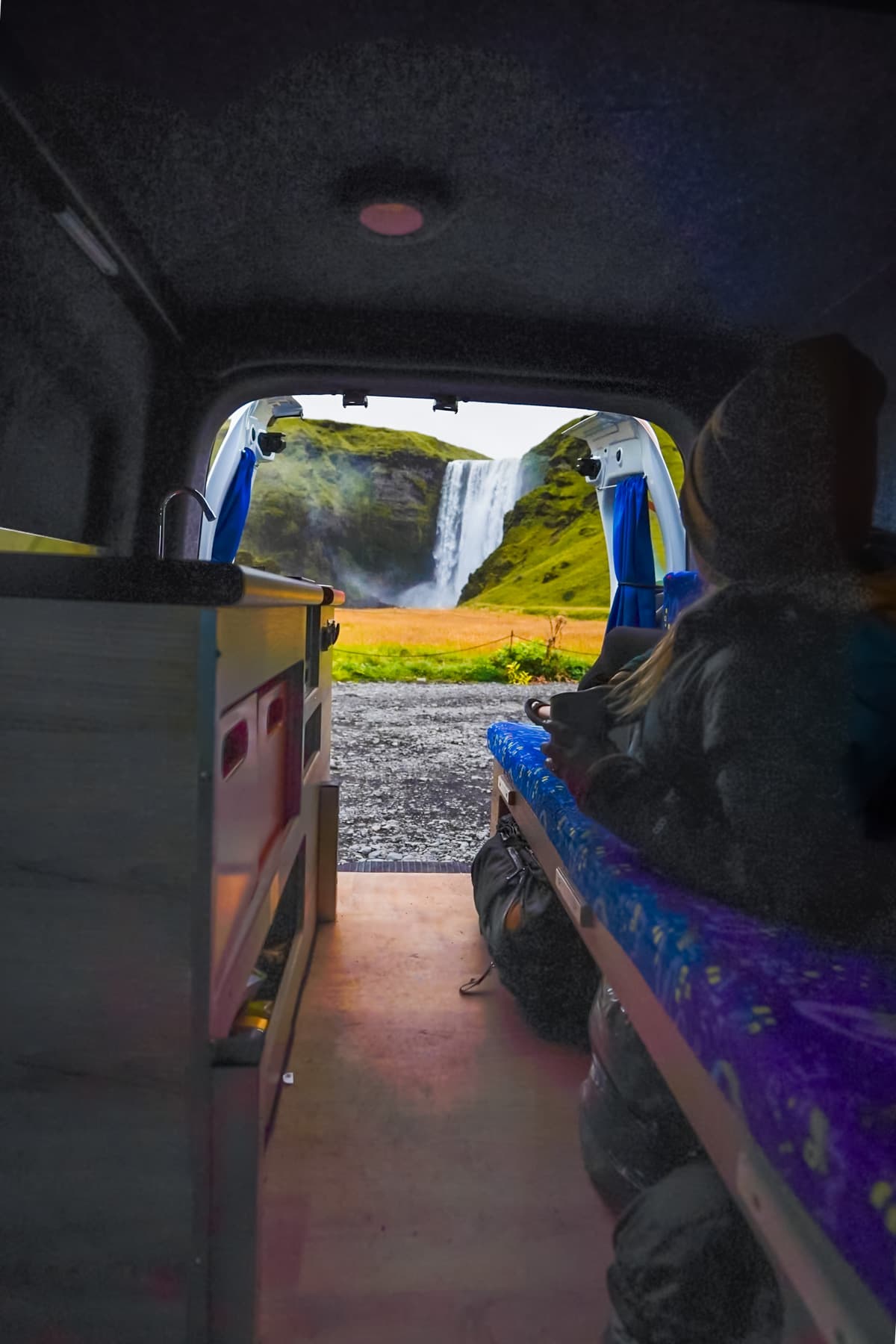 Woman laying on a couch in a campervan with open doors overlooking the Selfoos waterfall in Iceland