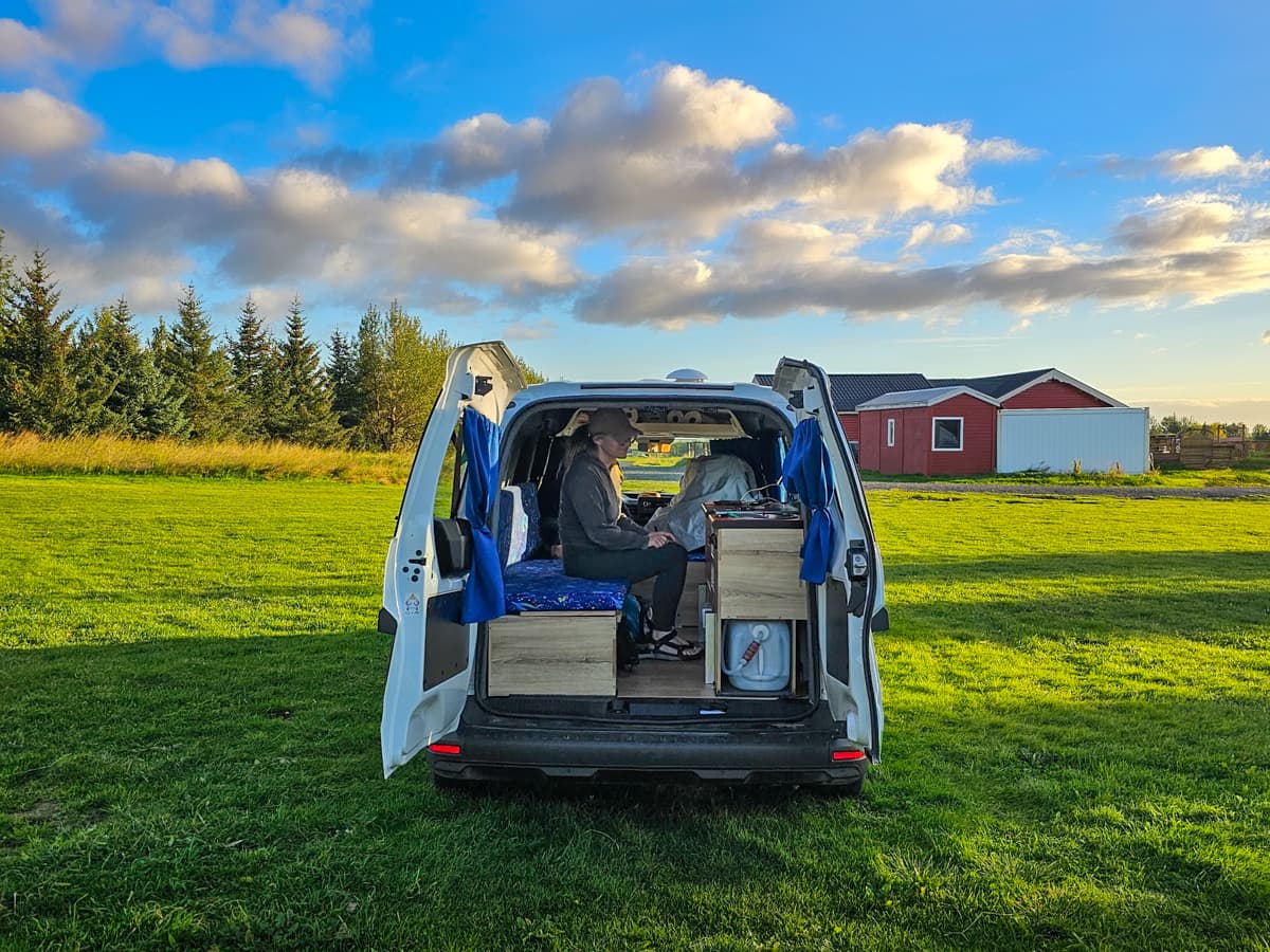 Woman sitting inside of a campervan in a grassy field with a red building in the background in Iceland
