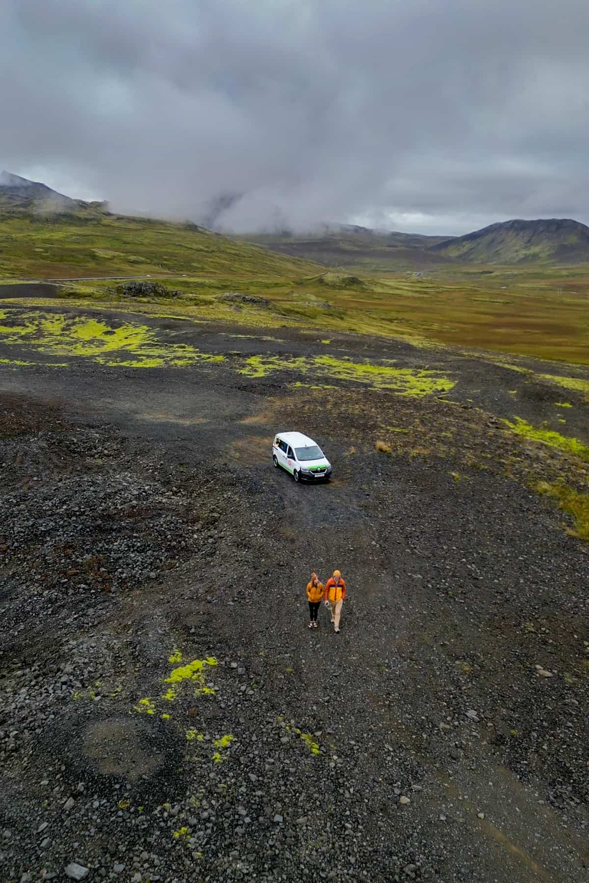 Couple walking away from their campervan while parked on a volcanic field with mossy green mountains in the background in Iceland