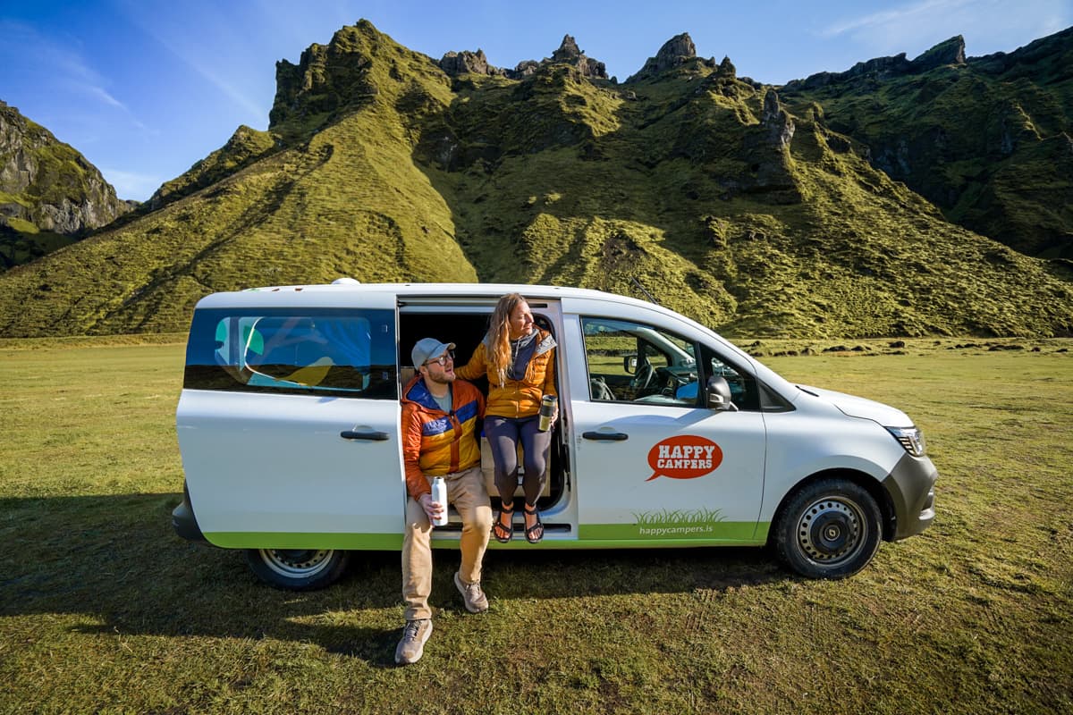 Couple sitting in the open door of a campervan with mossy green mountains in the background in the Pakgil campground of Iceland