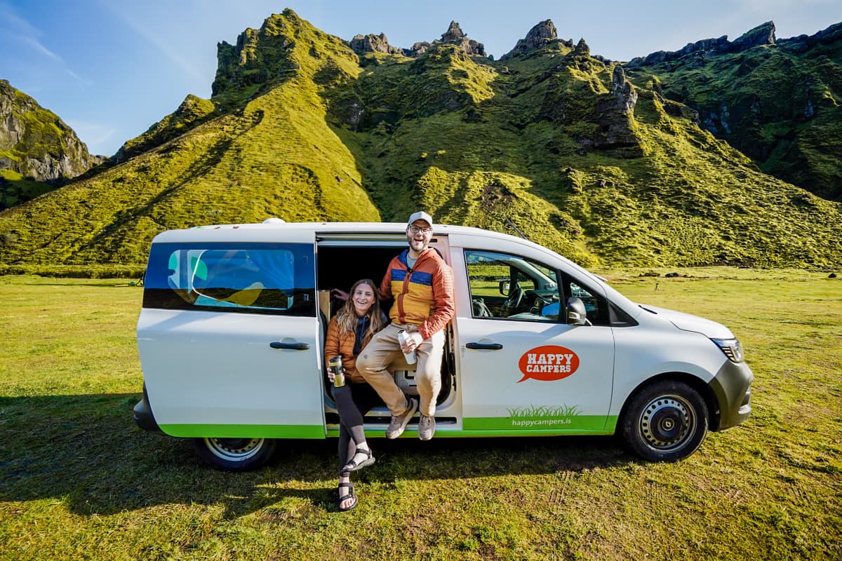 Couple sitting in the door of a campervan with rugged green mountains in the background in the Þakgil campground in the Highlands of Iceland