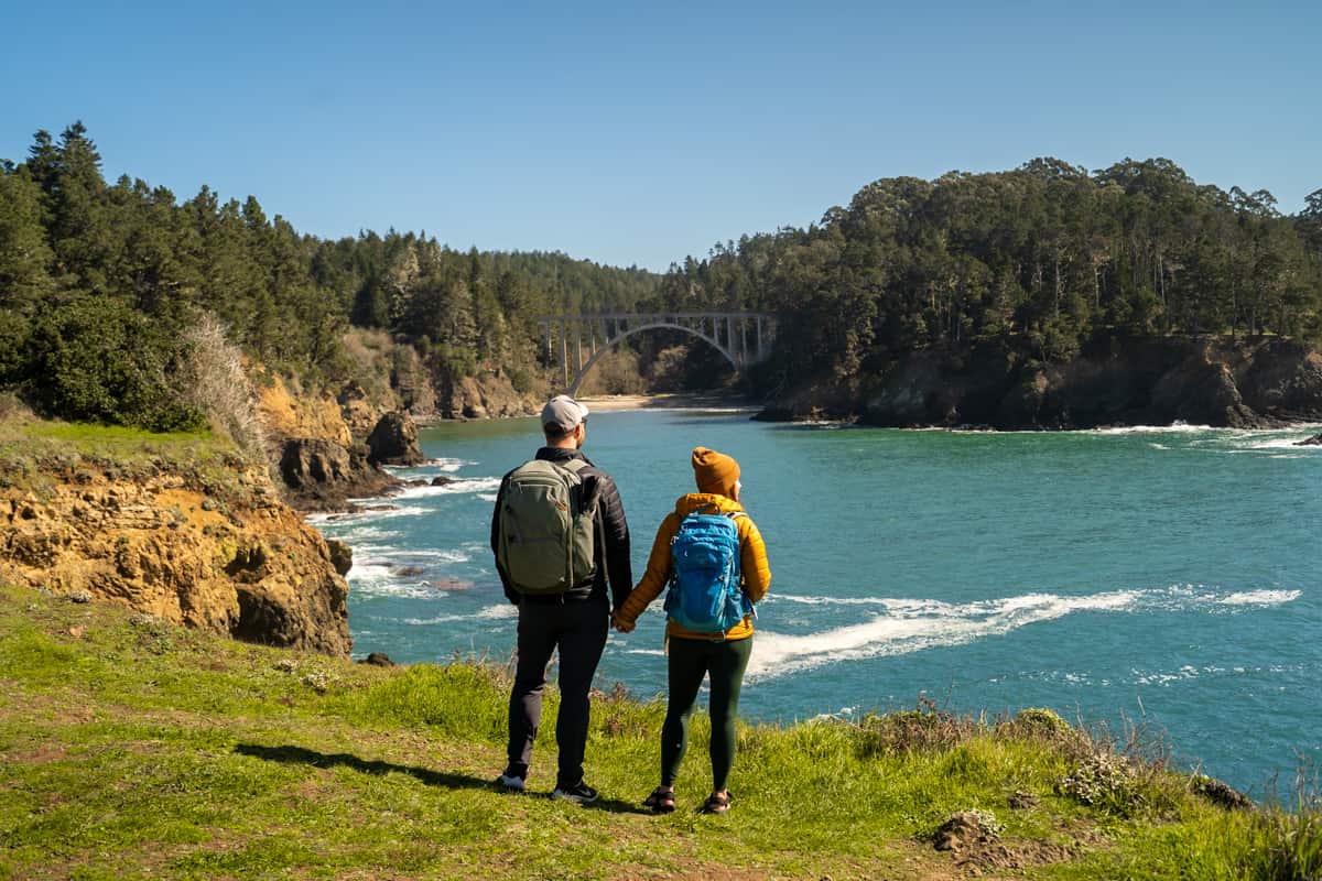 Couple holding hands along the Headlands Trail with a rugged bay and Russian Gulch Bridge in the background in Russian Gulch State Park in Mendocino, California