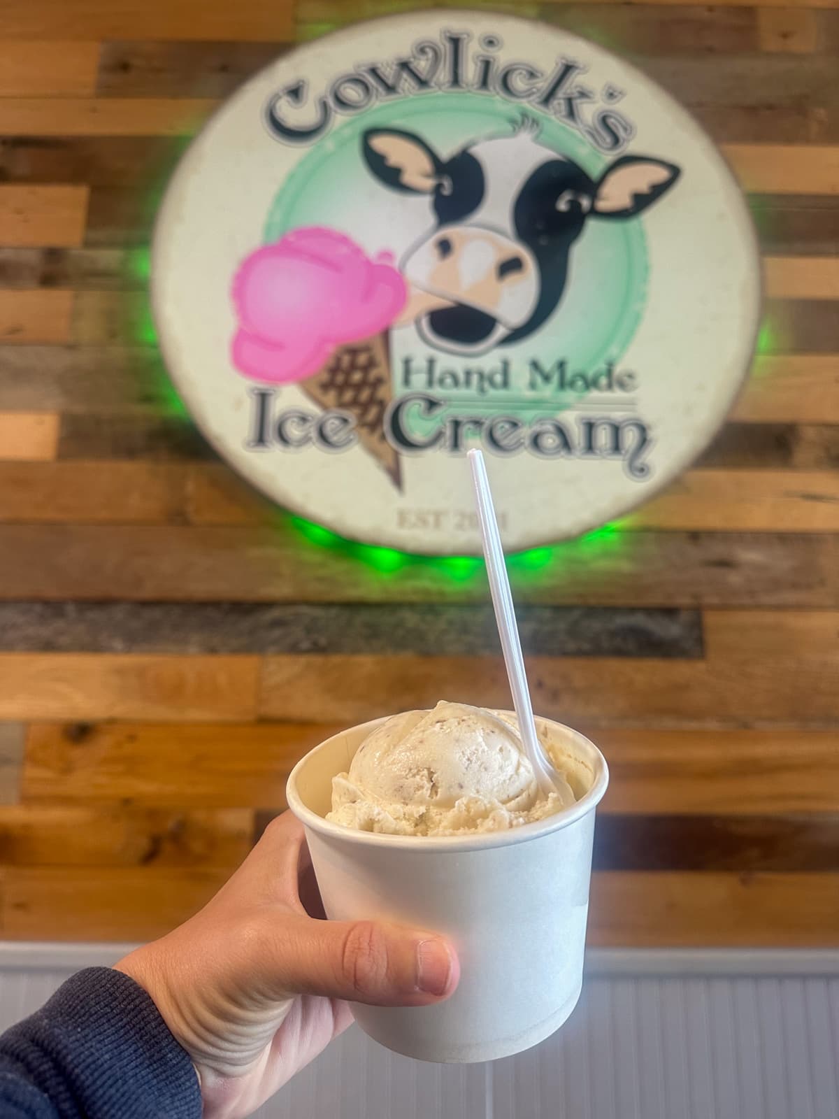 Cup of candy cap mushroom ice cream held in front of a Cowlick's Handmade Ice Cream sign in Fort Bragg, California