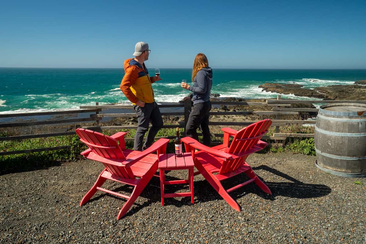 Couple standing by a fence in front of Adirondack chairs with rocky shorelines and the Pacific Ocean in the background at the Pacific Star Winery in Fort Bragg, California
