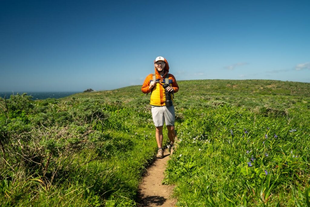 Man smiling on a trail surrounded by tall grasses and wildflowers on the Tomales Point Trail in Point Reyes National Seashore in California